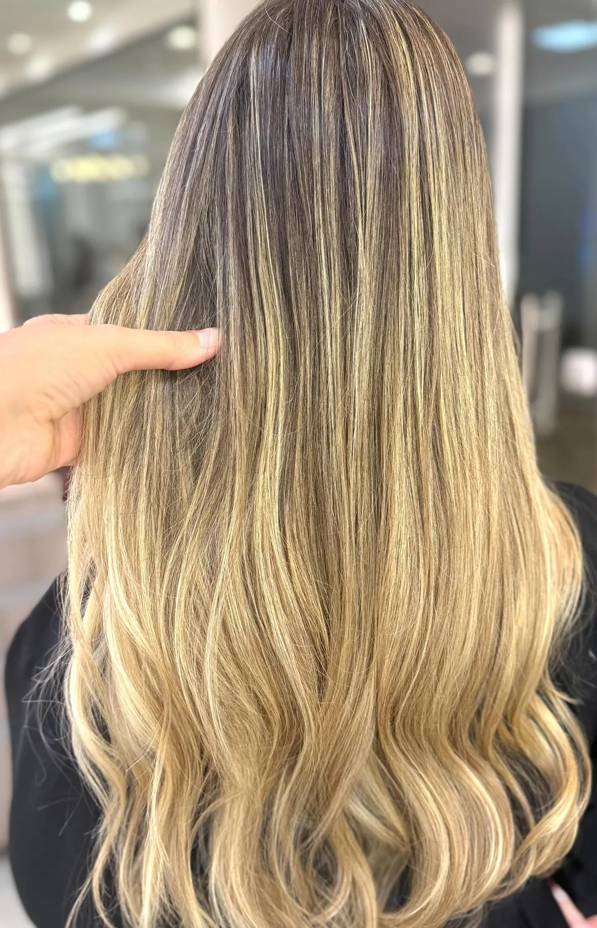 Cabelo longo de costas com mechas loiras iluminadas e raiz esfumada, segurado pela mão da cabeleireira
