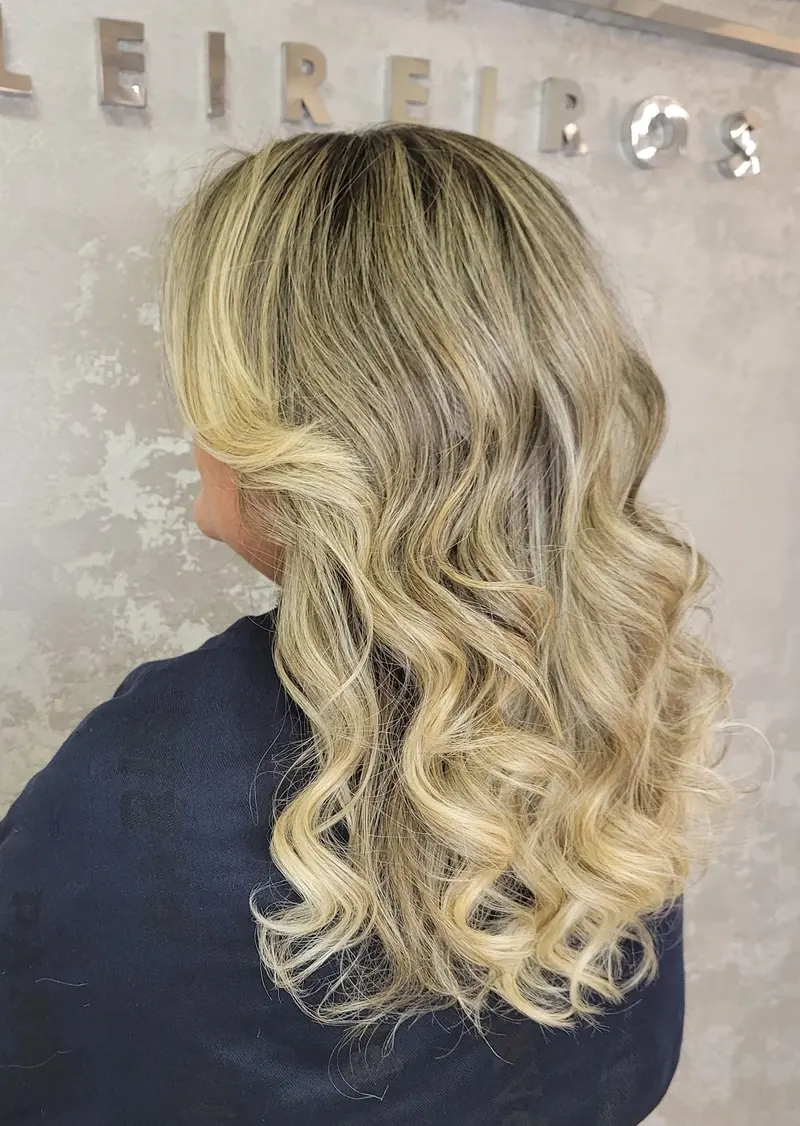 Cliente de costas com cabelo loiro em ondas, diante da parede com o nome do salão